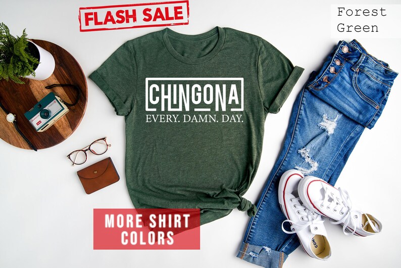 May include: A forest green t-shirt with the text "CHINGONA EVERY. DAMN. DAY." in white. The shirt is laid flat on a white surface with a pair of jeans, white sneakers, and other accessories.