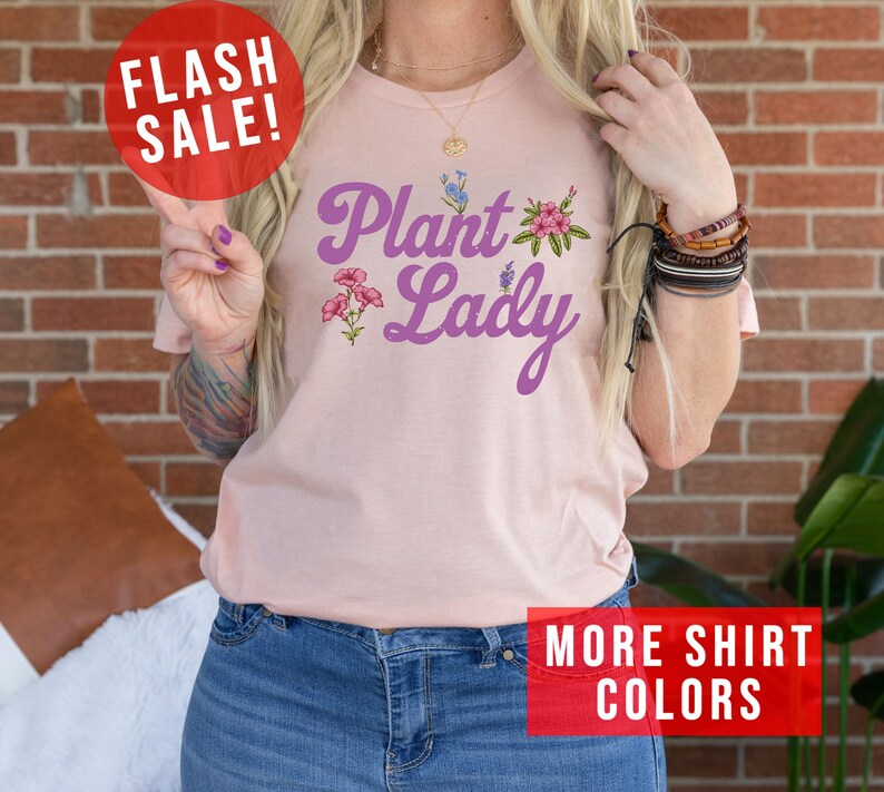 May include: A pink t-shirt with the text "Plant Lady" in purple script with floral accents. The shirt is being modeled by a person wearing blue jeans.