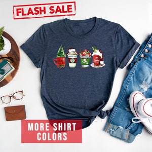 May include: Heather navy t-shirt featuring a festive design of four holiday-themed coffee cups. Each cup is decorated with seasonal elements like a Christmas tree, whipped cream, and holiday phrases. The shirt is paired with blue jeans and white sneakers.