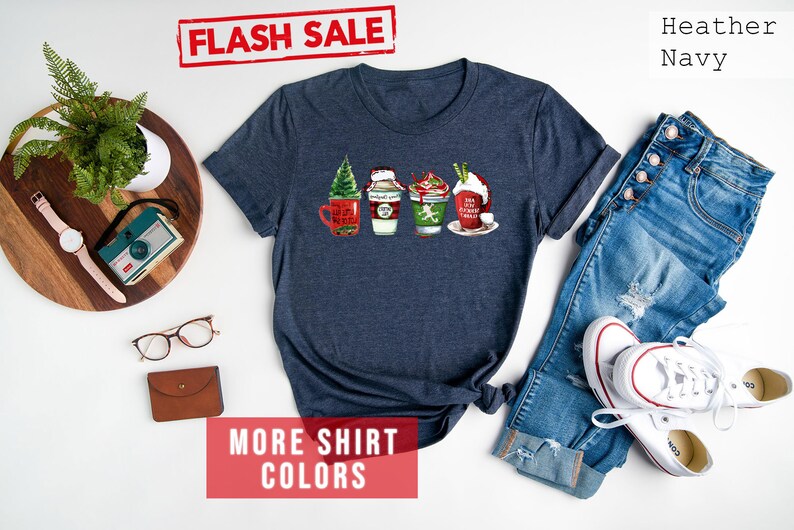 May include: Heather navy t-shirt featuring a festive design of four holiday-themed coffee cups. Each cup is decorated with seasonal elements like a Christmas tree, whipped cream, and holiday phrases. The shirt is paired with blue jeans and white sneakers.
