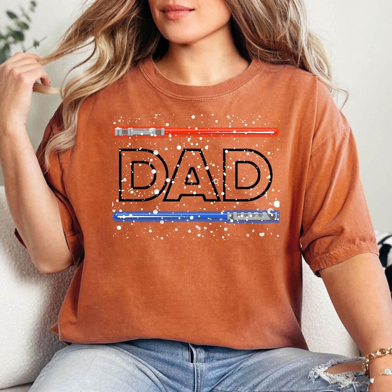 May include: Burnt orange t-shirt with the word "DAD" in black outlined letters. Above the word is a red lightsaber, and below is a blue lightsaber. White speckles are scattered around the text and lightsabers.