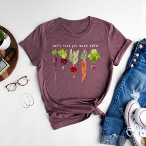 May include: Heather maroon t-shirt with a watercolor illustration of various vegetables and the text "Let's root for each other." The shirt is styled with blue jeans and white sneakers.