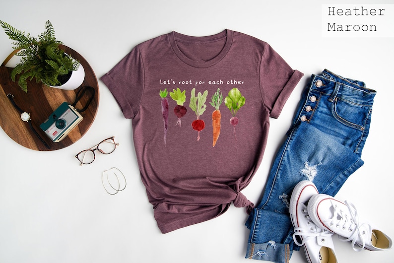 May include: Heather maroon t-shirt with a watercolor illustration of various vegetables and the text "Let's root for each other." The shirt is styled with blue jeans and white sneakers.