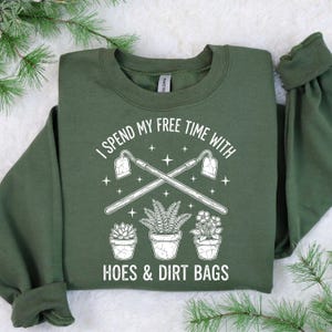 May include: Dark green sweatshirt featuring the phrase "I spend my free time with hoes & dirt bags." The design includes crossed gardening tools, potted plants, and stars. The sweatshirt is a Gildan brand.