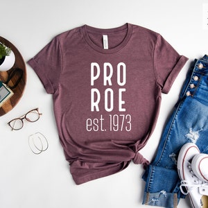 Op de afbeelding: Een heide-maron t-shirt met de tekst "PRO ROE est. 1973" in wit gedrukt. Het shirt ligt plat op een wit oppervlak met een spijkerbroek, een paar witte sneakers, een plant in een pot en een camera.