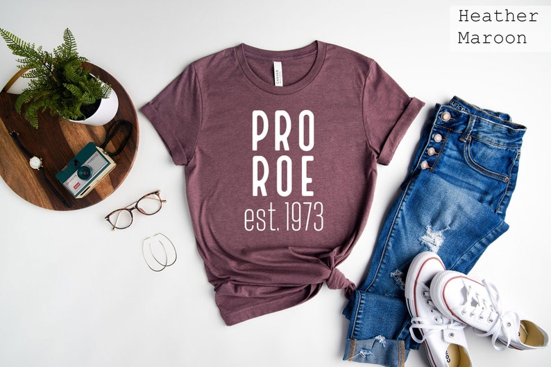 Op de afbeelding: Een heide-maron t-shirt met de tekst "PRO ROE est. 1973" in wit gedrukt. Het shirt ligt plat op een wit oppervlak met een spijkerbroek, een paar witte sneakers, een plant in een pot en een camera.