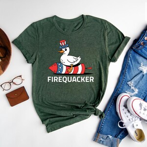 Peut inclure: T-shirt vert avec un dessin de canard portant un chapeau patriotique et chevauchant un feu d'artifice rouge, blanc et bleu. Le mot "FIREQUACKER" est imprim&eacute; en blanc. Le t-shirt est pr&eacute;sent&eacute; avec un jean bleu et des baskets blanches.