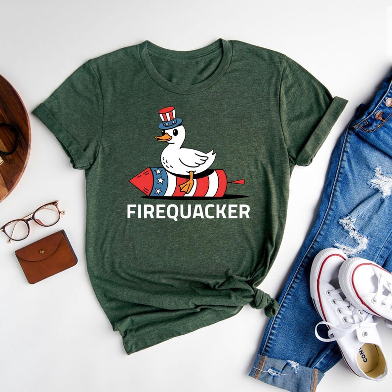 Peut inclure: T-shirt vert avec un dessin de canard portant un chapeau patriotique et chevauchant un feu d'artifice rouge, blanc et bleu. Le mot "FIREQUACKER" est imprim&eacute; en blanc. Le t-shirt est pr&eacute;sent&eacute; avec un jean bleu et des baskets blanches.