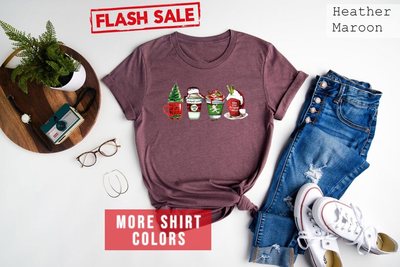 May include: Heather maroon t-shirt with a festive coffee cup design. The shirt features four different coffee cup designs with Christmas themes. The words "FLASH SALE" are in a red stamp. The words "MORE SHIRT COLORS" are in a red box.