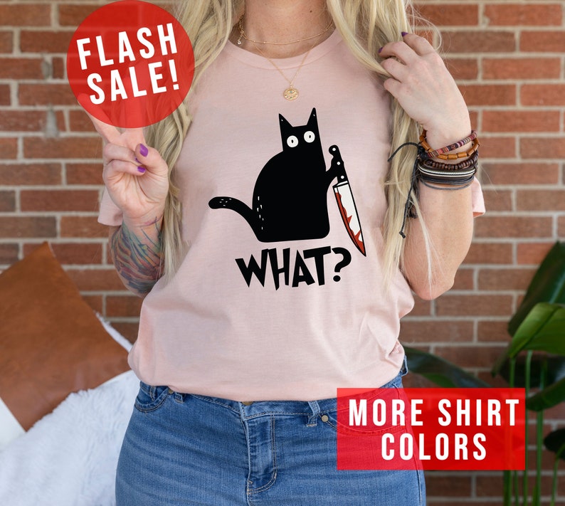 Op de afbeelding: Een roze T-shirt met een zwarte kat die een bloedig mes vasthoudt. De kat kijkt naar de kijker en de tekst "WHAT?" staat onder de kat gedrukt. Het shirt wordt gedragen door een persoon met lang blond haar.