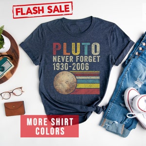 Op de afbeelding: Een marineblauw t-shirt met de tekst "Pluto Never Forget 1930-2006" en een retro-afbeelding van Pluto. Het shirt ligt op een wit oppervlak met een spijkerbroek, een paar witte sneakers, een houten dienblad met een plant, een camera, een horloge en een bruine portemonnee.