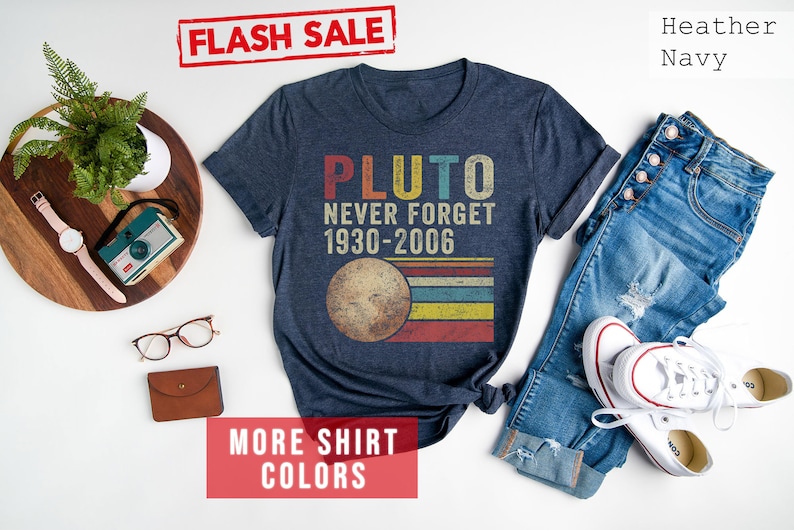 Op de afbeelding: Een marineblauw t-shirt met de tekst "Pluto Never Forget 1930-2006" en een retro-afbeelding van Pluto. Het shirt ligt op een wit oppervlak met een spijkerbroek, een paar witte sneakers, een houten dienblad met een plant, een camera, een horloge en een bruine portemonnee.