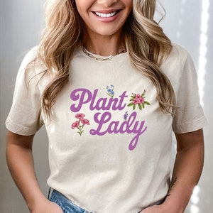 May include: A light beige t-shirt with a purple graphic that says "Plant Lady" with floral accents.