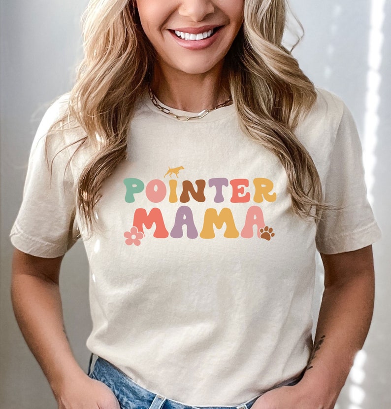 May include: A beige t-shirt with the text "POINTER MAMA" in colourful retro-style letters. A small silhouette of a dog is above the text, and a flower and paw print are below.