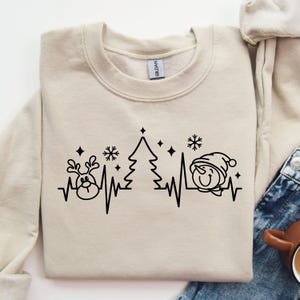 K&ouml;nnte beinhalten: Beiges Sweatshirt mit Rundhalsausschnitt und schwarzer Grafik eines Rentiers, Weihnachtsbaums und Schneemanns innerhalb einer Herzschlaglinie. Das Design umfasst Schneeflocken und Sterne und schafft ein festliches Urlaubsthema.