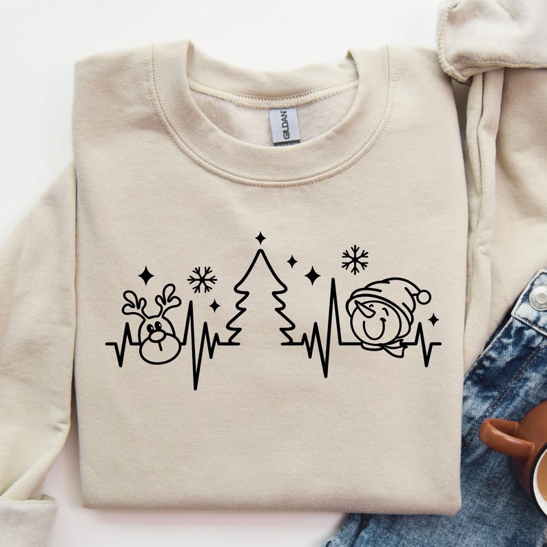 K&ouml;nnte beinhalten: Beiges Sweatshirt mit Rundhalsausschnitt und schwarzer Grafik eines Rentiers, Weihnachtsbaums und Schneemanns innerhalb einer Herzschlaglinie. Das Design umfasst Schneeflocken und Sterne und schafft ein festliches Urlaubsthema.