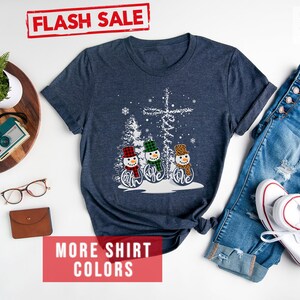 May include: Heather navy t-shirt featuring a snowman design with the word "believe". The snowmen wear hats and scarves in red, green, and leopard print. The shirt is paired with blue jeans and white sneakers.