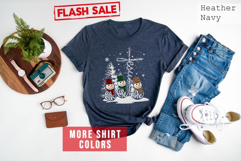 May include: Heather navy t-shirt featuring a snowman design with the word "believe". The snowmen wear hats and scarves in red, green, and leopard print. The shirt is paired with blue jeans and white sneakers.