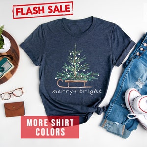May include: Heather navy t-shirt with a Christmas tree design on a sled, with the words "merry + bright". The shirt is paired with blue jeans and white sneakers. The words "FLASH SALE" are in a red box.