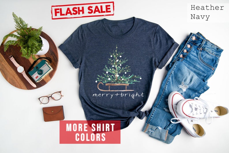 May include: Heather navy t-shirt with a Christmas tree design on a sled, with the words "merry + bright". The shirt is paired with blue jeans and white sneakers. The words "FLASH SALE" are in a red box.
