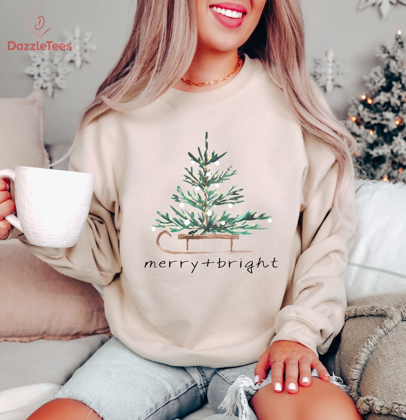 May include: Beige sweatshirt featuring a watercolor Christmas tree design on a sled, with the text "merry + bright" in black. The tree is green with white lights. The sweatshirt has a crew neck.