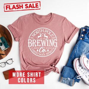 May include: Heather mauve t-shirt with a white circular graphic that reads "North Pole Brewing Co. Premium Christmas Spirits." The shirt is styled with blue jeans and white sneakers. A "Flash Sale" banner is visible.