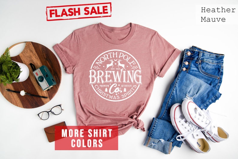 May include: Heather mauve t-shirt with a white circular graphic that reads "North Pole Brewing Co. Premium Christmas Spirits." The shirt is styled with blue jeans and white sneakers. A "Flash Sale" banner is visible.