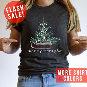 May include: Dark gray t-shirt featuring a watercolor Christmas tree on a sled, with the words "merry + bright" in white. A red circle with "FLASH SALE!" is in the upper left. The bottom right corner has a red rectangle with the words "MORE SHIRT COLORS".