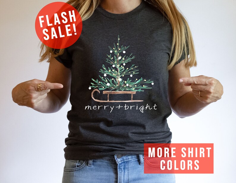 May include: Dark gray t-shirt featuring a watercolor Christmas tree on a sled, with the words "merry + bright" in white. A red circle with "FLASH SALE!" is in the upper left. The bottom right corner has a red rectangle with the words "MORE SHIRT COLORS".
