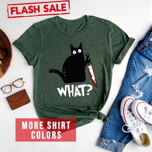 Op de afbeelding: Een bosgroen T-shirt met een zwarte kat die een bloedig mes vasthoudt en de tekst "WHAT?" erop gedrukt. Het shirt ligt op een wit oppervlak met een spijkerbroek, een paar witte sneakers en andere accessoires.