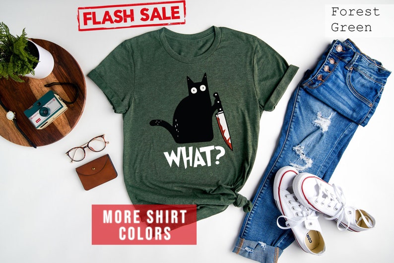 Op de afbeelding: Een bosgroen T-shirt met een zwarte kat die een bloedig mes vasthoudt en de tekst "WHAT?" erop gedrukt. Het shirt ligt op een wit oppervlak met een spijkerbroek, een paar witte sneakers en andere accessoires.