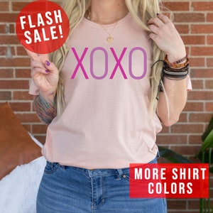 May include: A pink t-shirt with the word "XOXO" printed in pink and purple. The shirt is being modelled by a person wearing blue jeans.
