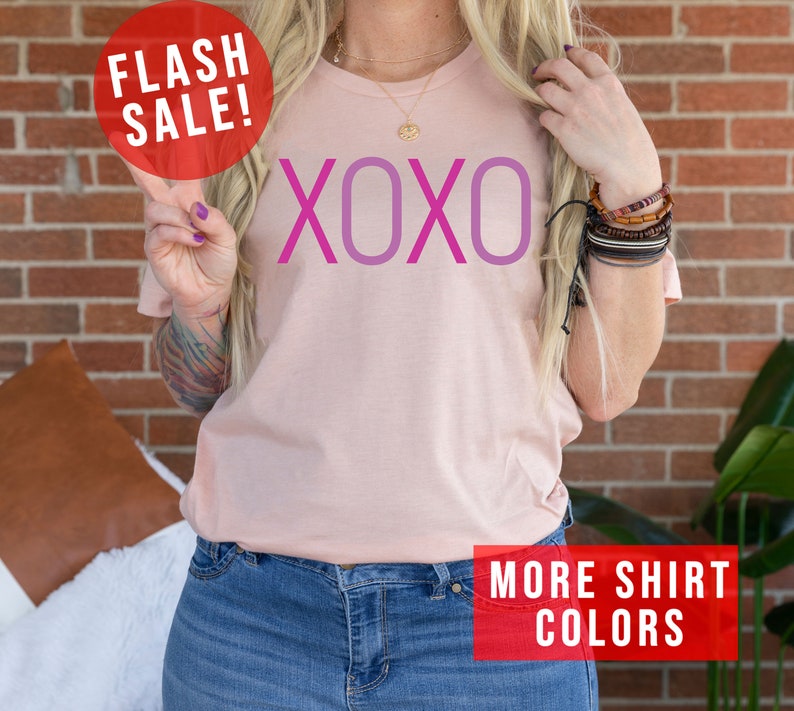 May include: A pink t-shirt with the word "XOXO" printed in pink and purple. The shirt is being modelled by a person wearing blue jeans.