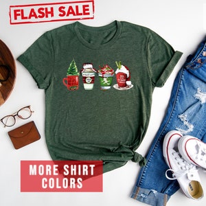 May include: Forest green t-shirt featuring a festive design of four holiday-themed beverages. The shirt has rolled-up sleeves and is paired with blue jeans and white sneakers. The words "FLASH SALE" are stamped in red.