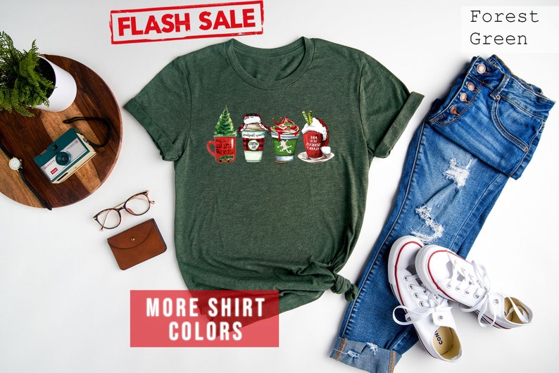 May include: Forest green t-shirt featuring a festive design of four holiday-themed beverages. The shirt has rolled-up sleeves and is paired with blue jeans and white sneakers. The words "FLASH SALE" are stamped in red.