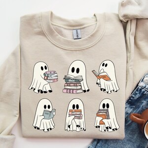 K&ouml;nnte beinhalten: Beiges Sweatshirt mit einer Grafik von sechs Cartoon-Geistern, die jeweils B&uuml;cher halten oder lesen. Die Geister sind wei&szlig; mit schwarzen Augen. Die B&uuml;cher sind in verschiedenen Farben, darunter Orange, Rosa und Blau. Das Sweatshirt hat einen Rundhalsausschnitt.