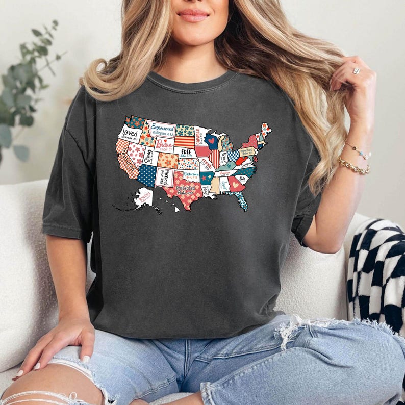 May include: A dark gray t-shirt with a patchwork design of the United States. Each state is filled with a different pattern and includes words like "Loved", "Strong", "Brave", "Free", and "Hope".