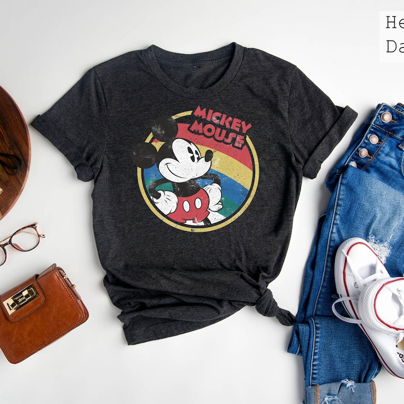 Puede incluir: Una camiseta gris oscuro con un gr&aacute;fico de estilo vintage de Mickey Mouse. El gr&aacute;fico presenta a Mickey Mouse con un atuendo rojo y amarillo, con un arco&iacute;ris detr&aacute;s de &eacute;l. El texto "Mickey Mouse" est&aacute; escrito en una fuente retro sobre Mickey Mouse.