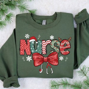 May include: A green Christmas sweatshirt with the word "Nurse" in festive, colorful letters. The letters are decorated with snowflakes, holly, and a red bow with a stethoscope.