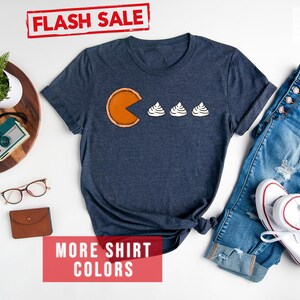 K&ouml;nnte beinhalten: Ein T-Shirt in Marineblau mit einem Aufdruck einer K&uuml;rbis-Tortenst&uuml;ckes mit einem Pac-Man-Mund, der drei wei&szlig;e Sahnekleckse frisst. Der Text "FLASH SALE!" ist in Rot auf wei&szlig;em Hintergrund &uuml;ber dem Shirt. Der Text "MORE SHIRT COLORS" ist in Wei&szlig; auf rotem Hintergrund unter dem Shirt.