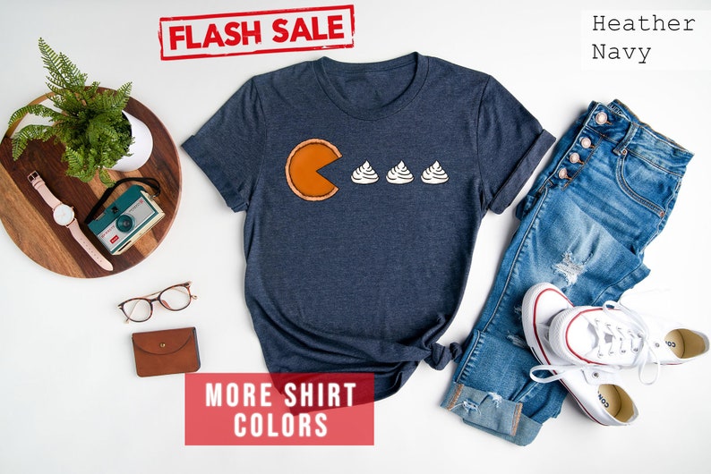 K&ouml;nnte beinhalten: Ein T-Shirt in Marineblau mit einem Aufdruck einer K&uuml;rbis-Tortenst&uuml;ckes mit einem Pac-Man-Mund, der drei wei&szlig;e Sahnekleckse frisst. Der Text "FLASH SALE!" ist in Rot auf wei&szlig;em Hintergrund &uuml;ber dem Shirt. Der Text "MORE SHIRT COLORS" ist in Wei&szlig; auf rotem Hintergrund unter dem Shirt.