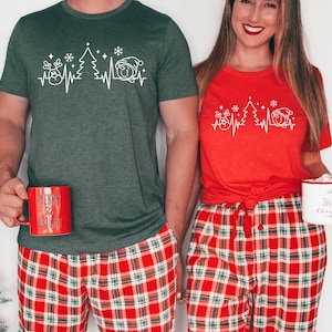 K&ouml;nnte beinhalten: Passende Weihnachtsshirts in Rot und Gr&uuml;n mit einem wei&szlig;en grafischen Herzschlag-Design mit Rentier, Weihnachtsbaum und Weihnachtsm&uuml;tze. Dazu rot-gr&uuml;ne karierte Schlafanzughosen. Jede Person h&auml;lt eine Tasse.