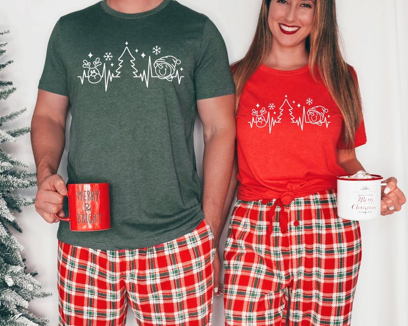 K&ouml;nnte beinhalten: Passende Weihnachtsshirts in Rot und Gr&uuml;n mit einem wei&szlig;en grafischen Herzschlag-Design mit Rentier, Weihnachtsbaum und Weihnachtsm&uuml;tze. Dazu rot-gr&uuml;ne karierte Schlafanzughosen. Jede Person h&auml;lt eine Tasse.