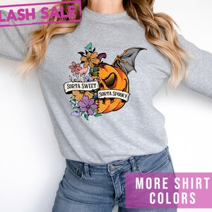 May include: A light gray sweatshirt featuring a Halloween-themed graphic. The graphic includes an orange pumpkin with bat wings, flowers, and the text "Sorta Sweet Sorta Spooky." The image also includes the text "Flash Sale" and "More Shirt Colors."