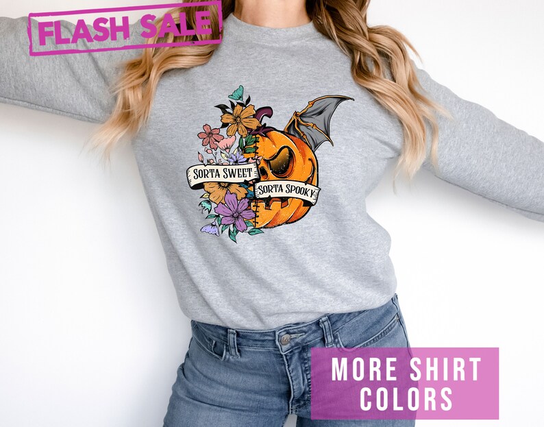 May include: A light gray sweatshirt featuring a Halloween-themed graphic. The graphic includes an orange pumpkin with bat wings, flowers, and the text "Sorta Sweet Sorta Spooky." The image also includes the text "Flash Sale" and "More Shirt Colors."