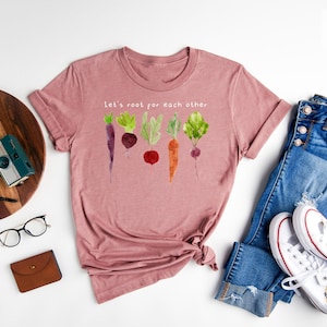 May include: Heather mauve t-shirt with the text "Let's root for each other" and a watercolor illustration of root vegetables. The shirt is paired with blue jeans and white sneakers.