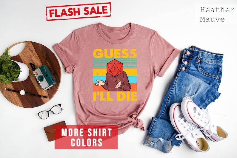 May include: A heather mauve t-shirt with a retro-style graphic of a person with a dice for a head. The text on the shirt reads "Guess I'll Die".