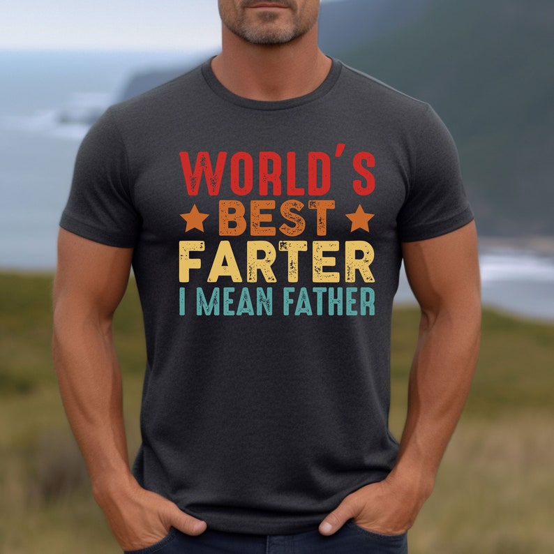 Puede incluir: Una camiseta gris oscuro con un gr&aacute;fico de estilo vintage que dice "World's Best Farter I Mean Father" en texto blanco, rojo, azul y amarillo.