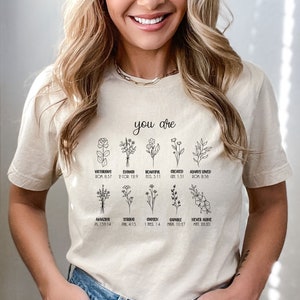 May include: A beige t-shirt with black line drawings of flowers and the text "you are" above the flowers. Each flower has a word and a bible verse underneath it. The words are: Victorious, Enough, Beautiful, Created, Always Loved, Amazing, Strong, Chosen, Capable, Never Alone.