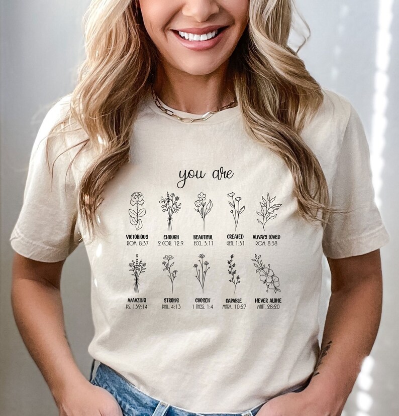 May include: A beige t-shirt with black line drawings of flowers and the text "you are" above the flowers. Each flower has a word and a bible verse underneath it. The words are: Victorious, Enough, Beautiful, Created, Always Loved, Amazing, Strong, Chosen, Capable, Never Alone.